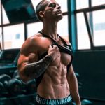 Muscular man showing abs and tattoos in a gym setting.